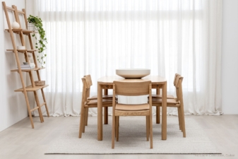 Scandinavian-inspired dining room with a natural oak table and four matching chairs with tan leather seats, styled with a minimalist bowl centerpiece and wooden ladder shelf against sheer white curtains.