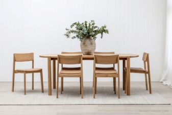 Oak dining table with six matching chairs and a centerpiece vase, in a bright, minimalist room.