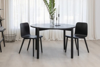 Modern black wooden dining table nz paired with two black chairs, styled with minimalist decor including a glass vase with greenery and tall candles, set against sheer curtains in a bright interior. Cuchi new Zealand
