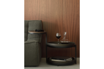 Modern round side table with black frame and dark trays, placed beside a green armchair and styled with a wine decanter, glass of red wine, and books against a wooden wall backdrop.