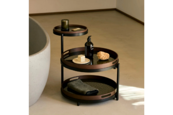 Three-tier round side table with black metal frame and dark wood trays, styled with bath accessories and placed beside a concrete bathtub in a minimalist bathroom setting.