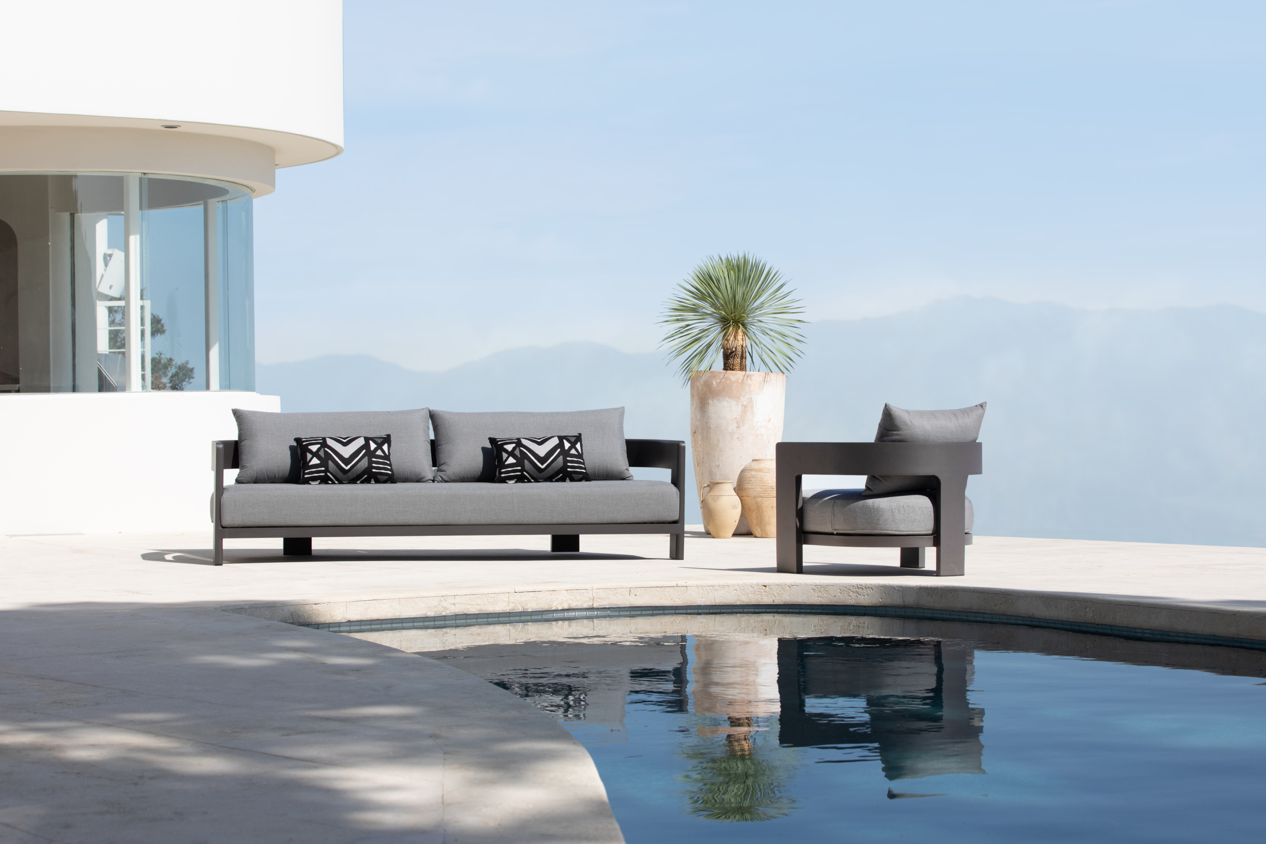 Modern outdoor patio with a grey sofa set, geometric black cushions, and a lounge chair beside a curved swimming pool, set against a mountain backdrop under a clear blue sky.